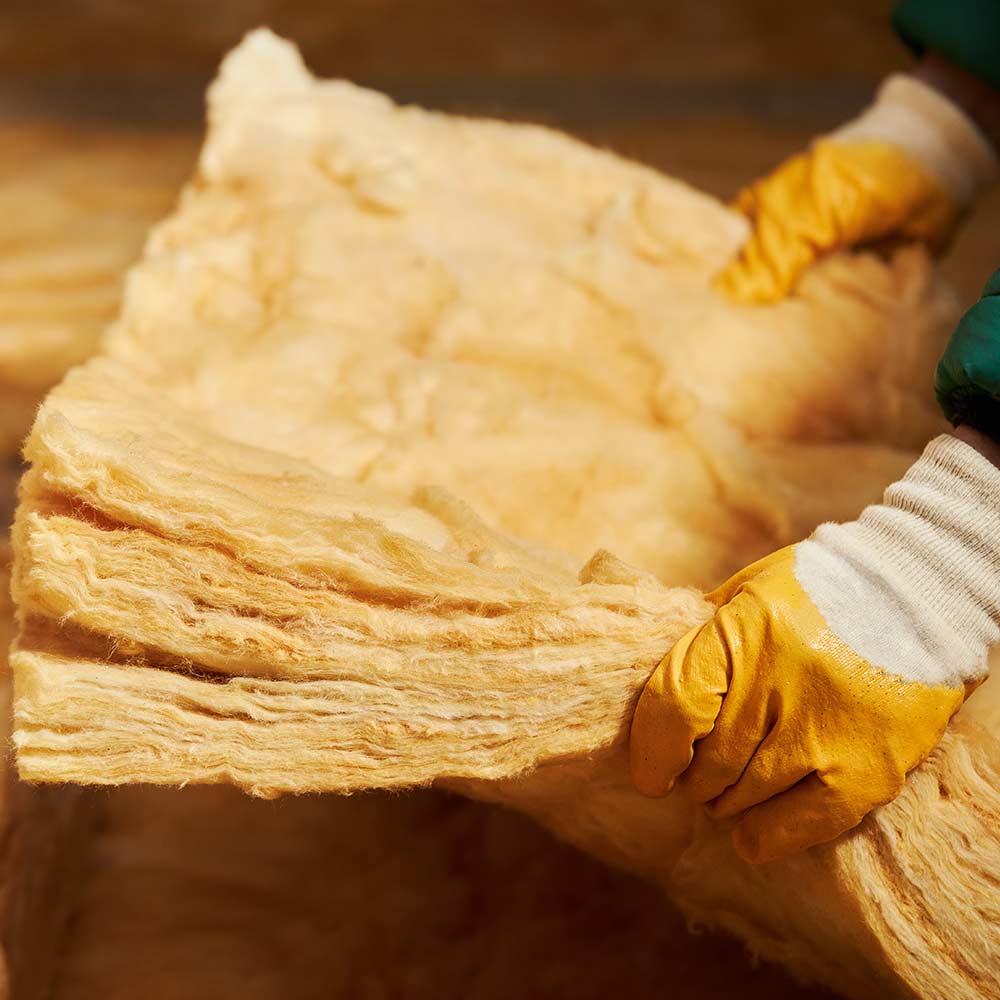 Isolation thermique : pose de laine de verre par un ouvrier. Mains gantées soulevant un rouleau épais de laine de verre jaune pour l'isolation thermique d'un bâtiment.