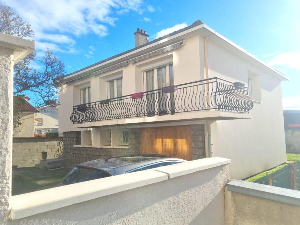 Maison blanche avec balcon noir, garage en bois et ciel bleu.