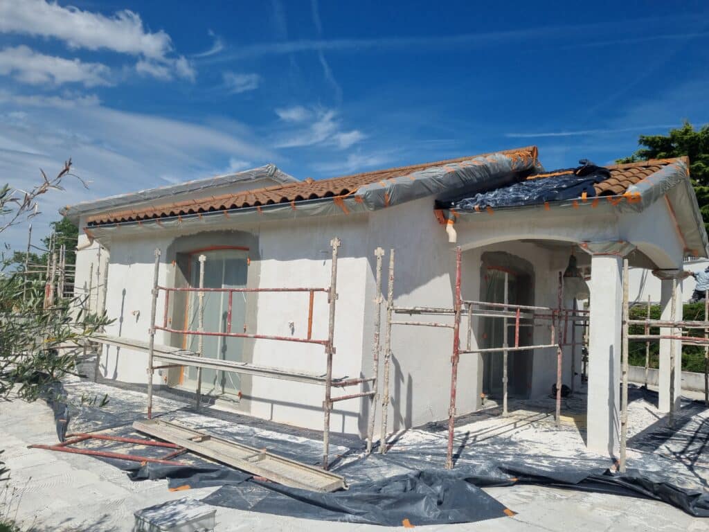 Maison blanche en rénovation avec échafaudage, toit en tuiles et bâches protectrices sous un ciel bleu ensoleillé.